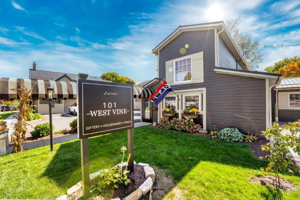 A charming retail shop with a sign reading "101 West Vine" surrounded by greenery and outdoor decor.