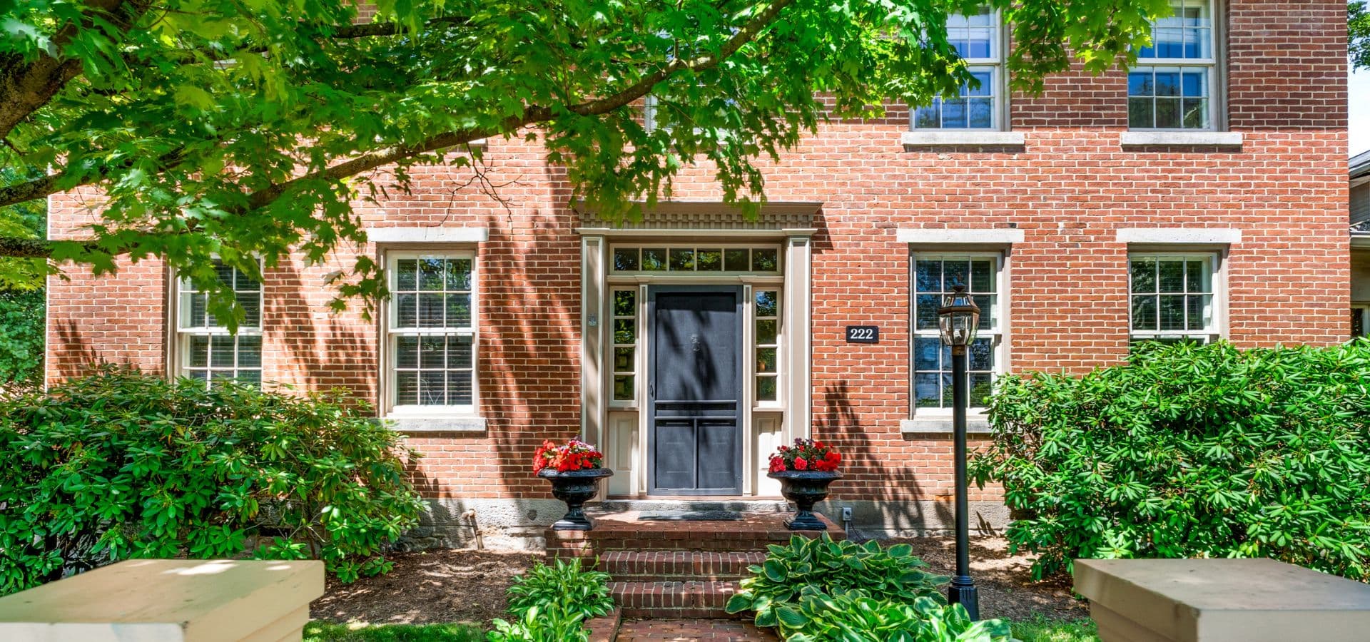 A brick house with a black front door, surrounded by greenery and flowers.