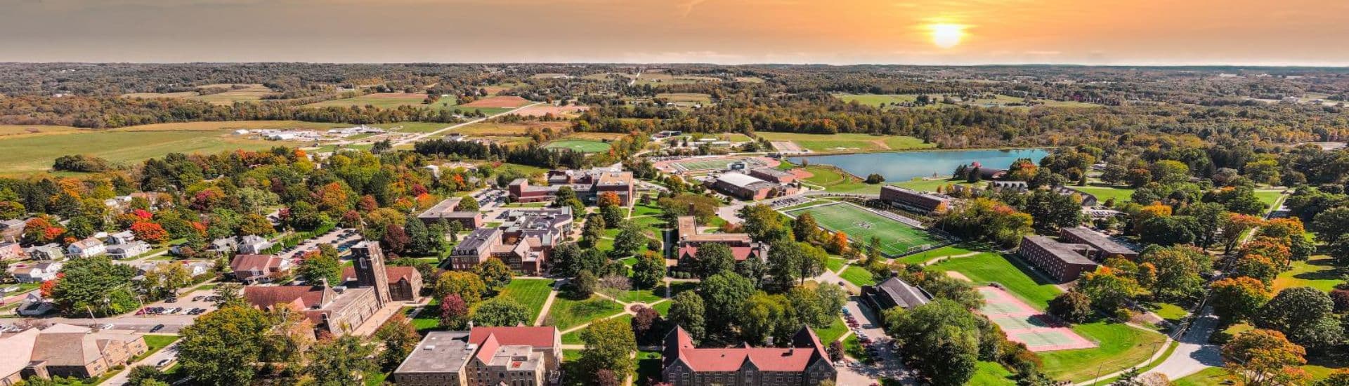 Aerial view of a college campus surrounded by colorful autumn trees and green fields at sunset.