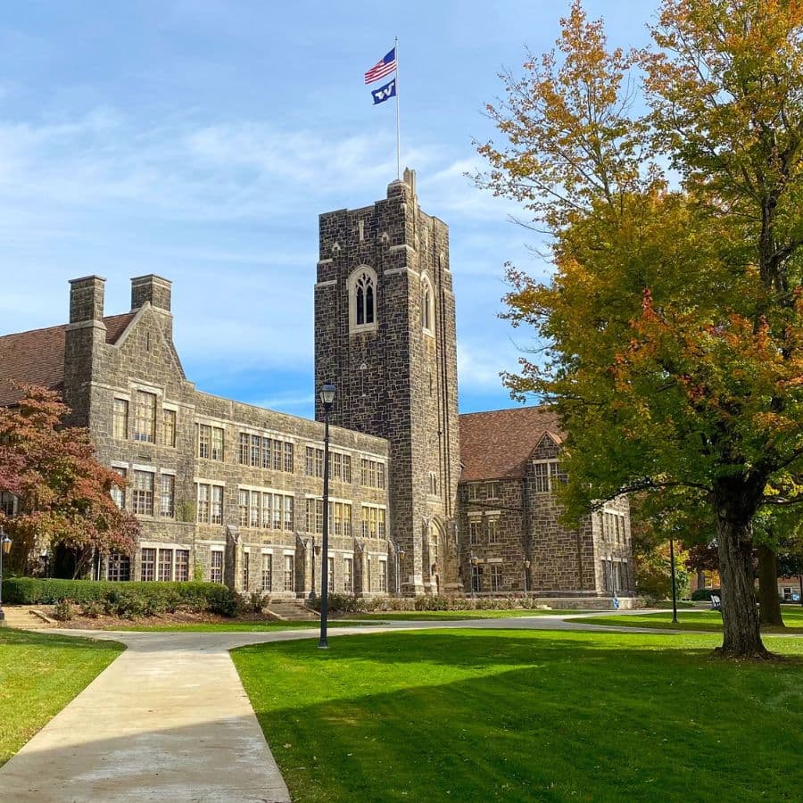 Historic stone building with a tall clock tower and American flag, surrounded by green grass and trees.